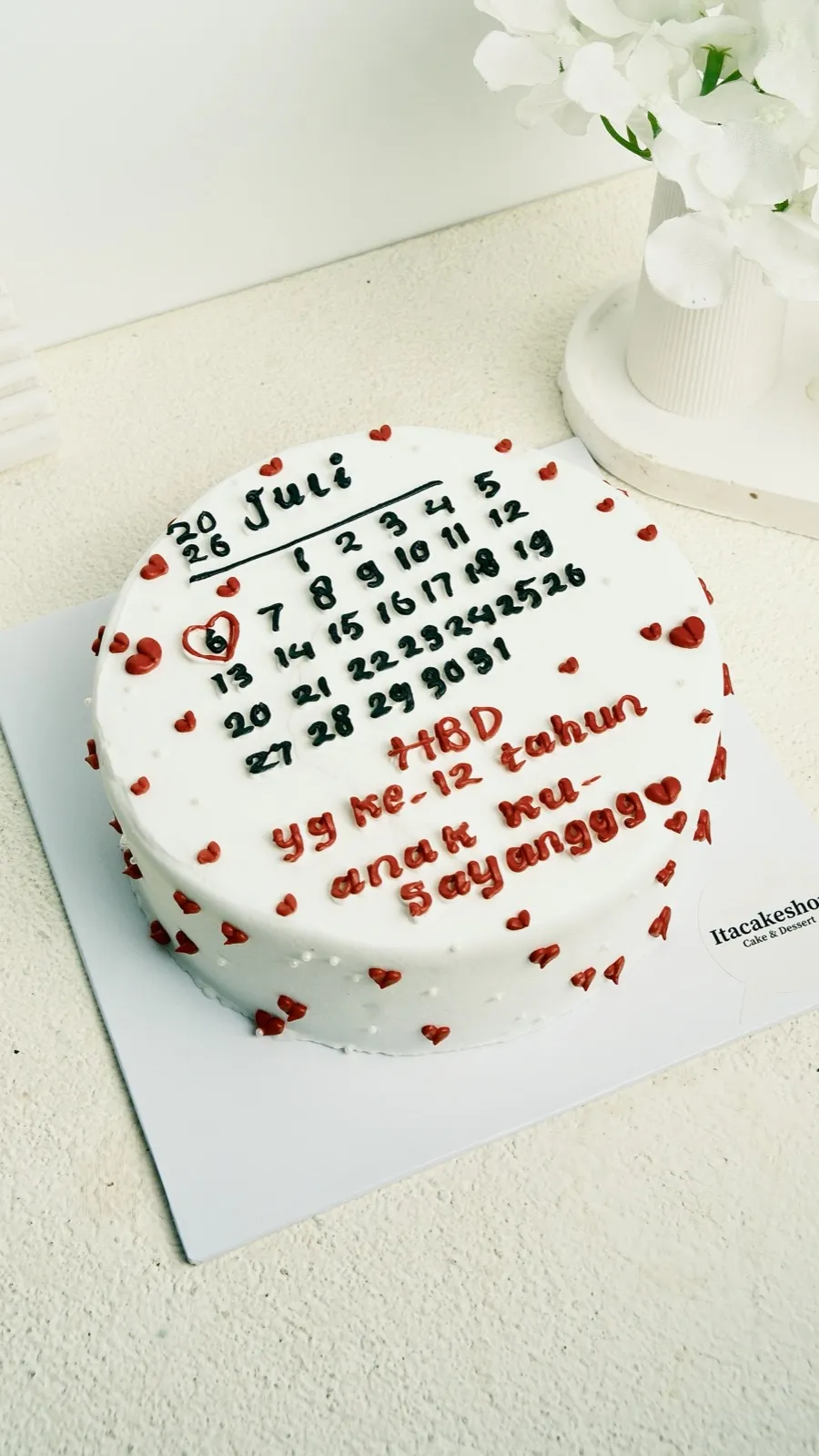 Cake 18 × 6 krim putih bertabur hati merah dengan tulisan kalender ulang tahun